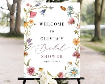 May include: A white welcome sign with a floral wreath border. The sign reads "Welcome to Olivia's Bridal Shower August 22, 2023".