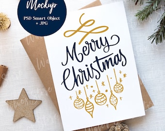 May include: A white greeting card with the words "Merry Christmas" in navy blue script and gold accents. The card is on a brown envelope, surrounded by gold stars, pine cones, and cinnamon sticks, creating a festive holiday theme.