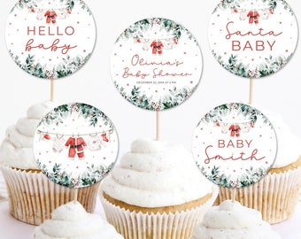 Könnte beinhalten: Cupcakes mit weißem Zuckerguss und dekorativen Aufsätzen. Die Aufsätze zeigen festliche Designs mit Texten wie "Hello Baby" und "Santa Baby", sowie Babykleidung und Grünzeug. Ideal für eine Babyparty.
