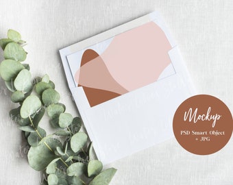 Op de afbeelding: Een witte envelop met een abstract ontwerp in roze en bruin aan de voorkant. De envelop ligt op een witte linnen stof met een takje eucalyptusbladeren links. Een bruine cirkel met de tekst "Mockup PSD Smart Object + JPG" bevindt zich in de rechterbenedenhoek.