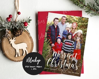 May include: A Christmas card mockup with a photo of a family in a forest setting. The card features the text "Merriest Christmas" in a white cursive font. The family is dressed in winter clothing and smiling. The card is on a white background with a wooden ornament and a red envelope.