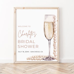 May include: A white sign with a wooden frame, featuring a champagne flute with a pearl necklace draped over it. The text reads "Welcome to Charlotte's Bridal Shower, July 18, 20XX - San Diego, CA".