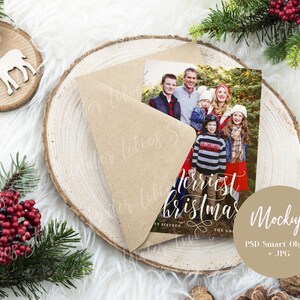 May include: A Christmas card mockup with a brown envelope and a photo of a family in front of a Christmas tree. The card reads "Merriest Christmas" in white script. The mockup is on a wooden background with a brown circle that says "Mockup PSD Smart Object + JPG".