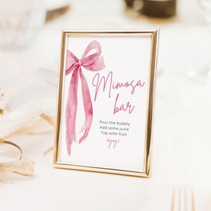 May include: A gold-framed sign with a pink watercolor bow and the text "Mimosa Bar" with instructions to "Pour the bubbly, Add some juice, Top with fruit, enjoy!"