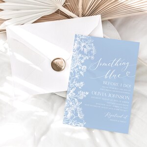 May include: A light blue invitation card with white floral designs and the text "Something Blue Before I Do!"  Olivia Johnson Saturday, August 22, 2023 at 4:00PM  Katherine's Garden, 123 Main Street  RSVP by July 15th to [phone number]  Registered at [website]
