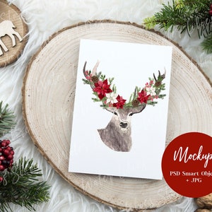 May include: A watercolor illustration of a deer with a floral crown made of red and white flowers. The illustration is on a white card, which is sitting on a wooden slice. The card is surrounded by greenery and a red circle with the text "Mockup PSD Smart Object + JPG".
