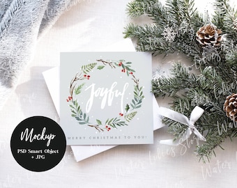 May include: A white card with a watercolor wreath of green leaves and red berries. The word "Joyful" is written in white script inside the wreath. The card has the text "Merry Christmas to You!" printed below the wreath.