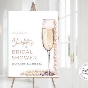 May include: A white welcome sign for a bridal shower with the text "Welcome to Charlotte's Bridal Shower" and the date "July 18, 20XX" and location "San Diego, CA". The sign features a champagne flute with a pearl necklace draped over it.