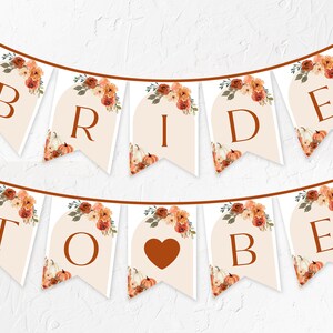 Puede incluir: Una guirnalda de banderines con la frase "BRIDE TO BE". Cada banderín tiene un fondo crema, letras marrones y está decorado con arreglos florales y calabazas con temática otoñal. Un corazón marrón reemplaza la "I" en "BRIDE".