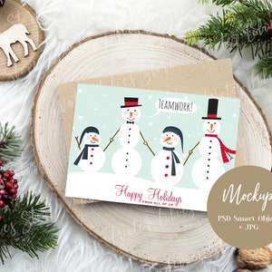 May include: A Christmas card with a light blue background featuring four snowmen holding hands. The snowmen are wearing hats, scarves, and buttons. The card says "Happy Holidays from all of us" and "Teamwork!" in red text.