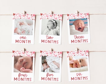 May include: Six photos of a baby girl, each photo is in a white frame with a pink and white heart pattern border. The photos are hung on a string with clothespins. Each photo has a number and the word "months" written below the photo. The photos show the baby at one, two, three, four, five, and six months old.
