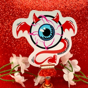 May include: A white sticker with a blue eye with pink veins and red devil horns and tail. The sticker has the text "@STICKYDICKDESIGN" on the side.
