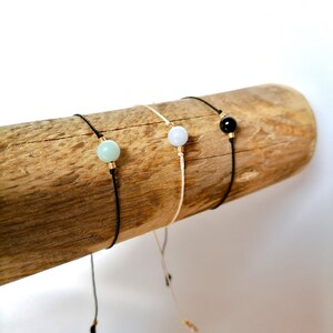 Peut inclure: Trois bracelets minimalistes avec des perles de pierres précieuses. L'un a une perle bleu clair sur un cordon noir, un autre a une perle bleu clair sur un cordon crème, et le troisième a une perle noire sur un cordon noir. Chaque bracelet a un accent doré.