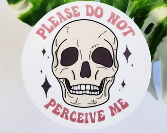 Peut inclure: Un autocollant rond blanc avec un crâne rose et le texte "Please Do Not Perceive Me" en lettres roses.