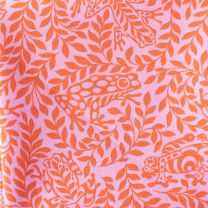 May include: Pink fabric with a repeating pattern of orange frogs and leaves. The frogs are stylized and have spots. The leaves are arranged in a flowing pattern.