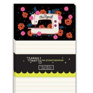 May include: A rectangular notepad with cream-colored lined paper. The top section features a black background with pink and orange flowers and a white sewing machine illustration with the text my therapist. A black band displays Sarah Watts for Craftedmoon and moda fabrics + supplies.