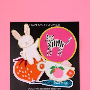 May include: Iron-on patches featuring a white bunny, a striped cat, a strawberry, a teacup with an orange, and a teal tool with the text "tear it up". The patches are on a black background with the text "IRON-ON PATCHES" and "www.renaissanceribbons.com".
