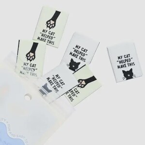May include: A collection of woven labels with the text "MY CAT 'HELPED' MAKE THIS" in black. The labels feature a black cat paw print or a cat silhouette. The labels are white and are in a clear plastic bag.