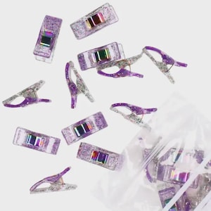 May include: A collection of purple glitter sewing clips with iridescent metal springs, some scattered and others contained in a clear plastic bag labeled with the text MORE ME KNOW GLITTER CLIPS MOREMEKNOW.COM, featuring a sparkly finish.
