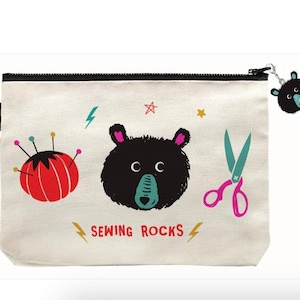 May include: Cream-colored sewing pouch with a black zipper and handle. The pouch features a cartoon bear face, a red pincushion, and teal scissors. Text on the pouch reads "SEWING ROCKS". A small bear charm hangs from the zipper.