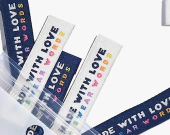 May include: A close-up of a package containing several blue and white fabric labels with the text "Made with Love + Swear Words" printed in rainbow colours. The labels are in a clear plastic bag with a white background.