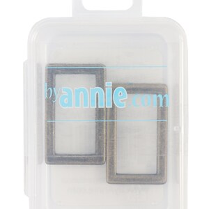 May include: Two antique bronze rectangular metal rings are displayed inside a clear plastic container. The container has a hanging tab at the top and the text "byannie.com" is visible on the container.