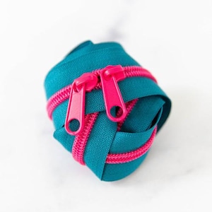 May include: A teal zipper is coiled, with a bright pink zipper pull. The zipper tape is teal, and the zipper teeth are pink. The zipper is on a white background.