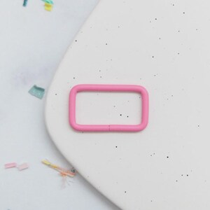 May include: A bright pink rectangular metal buckle. The buckle is a solid color and has rounded corners. The buckle is on a white surface with small black speckles and colorful confetti.