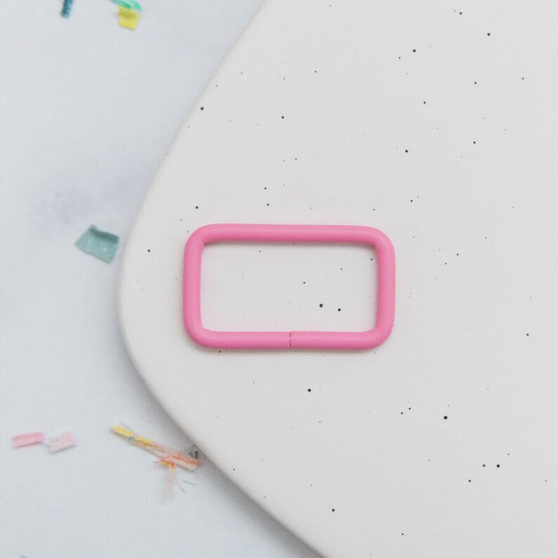 May include: A bright pink rectangular metal buckle. The buckle is a solid color and has rounded corners. The buckle is on a white surface with small black speckles and colorful confetti.