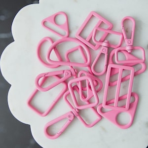 May include: A collection of pink metal hardware pieces, including D-rings, rectangular buckles, and swivel clasps. The hardware is arranged on a white, scalloped-edge surface. The pieces are all the same shade of bright pink.
