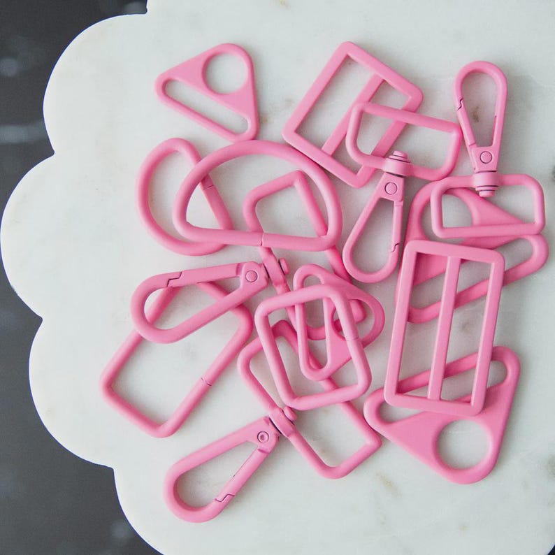 May include: A collection of pink metal hardware pieces, including D-rings, rectangular buckles, and swivel clasps. The hardware is arranged on a white, scalloped-edge surface. The pieces are all the same shade of bright pink.