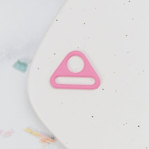 May include: A bright pink, plastic triangle ring with a circular hole at the top and a rectangular slot at the bottom. The ring is set against a white background with small black specks.