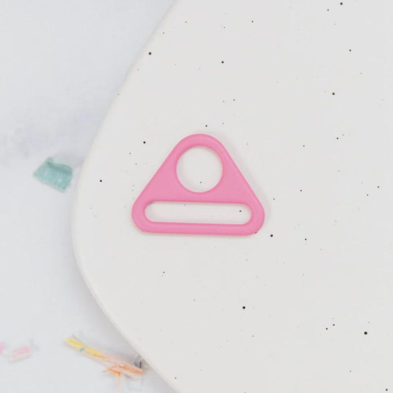 May include: A bright pink, plastic triangle ring with a circular hole at the top and a rectangular slot at the bottom. The ring is set against a white background with small black specks.