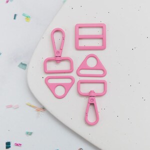 May include: A collection of five pink metal hardware pieces, including a swivel clasp, a rectangular slider with two bars, a triangle-shaped ring, and a D-ring with a swivel clasp. The hardware is arranged on a white surface with colorful confetti.