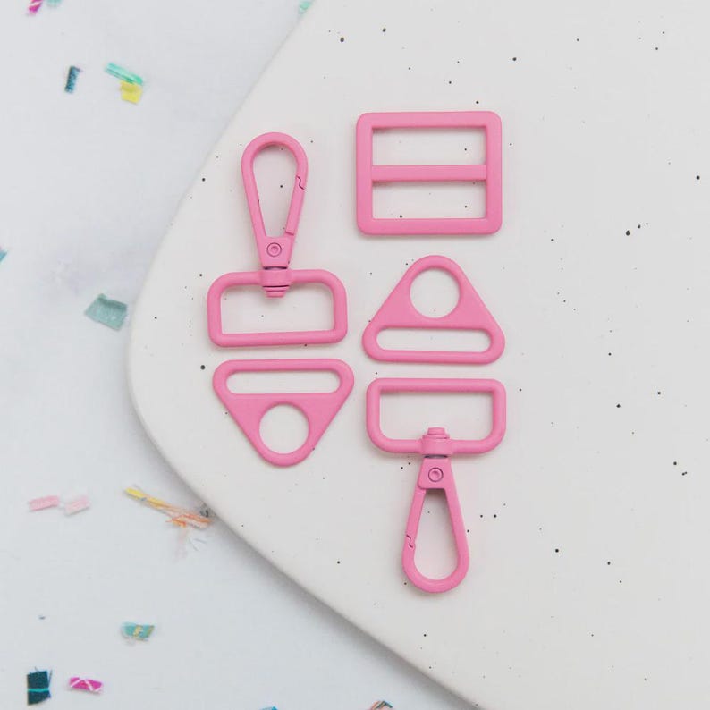 May include: A collection of five pink metal hardware pieces, including a swivel clasp, a rectangular slider with two bars, a triangle-shaped ring, and a D-ring with a swivel clasp. The hardware is arranged on a white surface with colorful confetti.