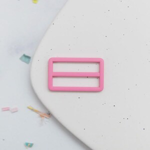 May include: A bright pink rectangular slider buckle with two horizontal bars. The buckle is made of plastic and is set against a white background with small black specks and colorful confetti.