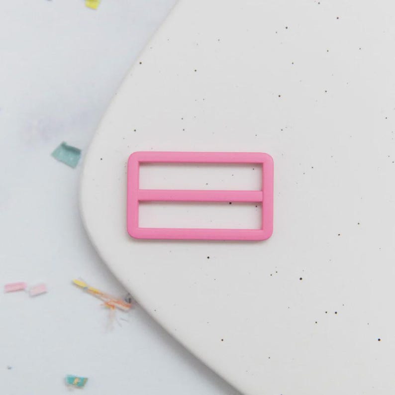 May include: A bright pink rectangular slider buckle with two horizontal bars. The buckle is made of plastic and is set against a white background with small black specks and colorful confetti.