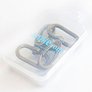 May include: A clear plastic container holding several antique bronze-colored swivel clasps. The clasps are oval-shaped and appear to be made of metal. The container has the text "annie.com" printed on it.