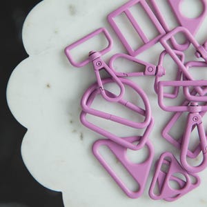 May include: A collection of lavender-colored metal hardware pieces, including D-rings, rectangular sliders, and swivel clasps. The hardware is arranged on a white, scalloped surface, with a dark background. These pieces are likely for bag making or other craft projects.