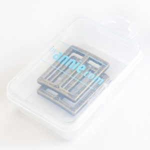 May include: Two antique brass-colored rectangular buckles are inside a clear plastic container. The buckles have a textured surface and a slotted design. The container has the text "byannie.com" printed on it in light blue.
