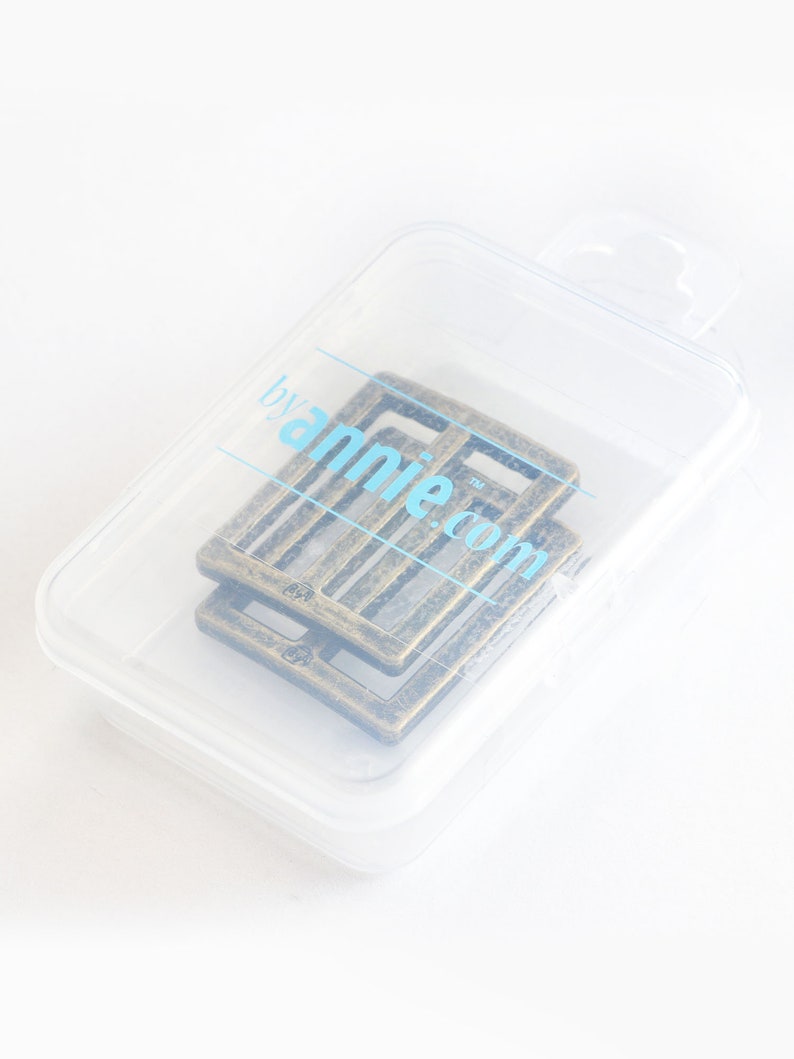 May include: Two antique brass-colored rectangular buckles are inside a clear plastic container. The buckles have a textured surface and a slotted design. The container has the text "byannie.com" printed on it in light blue.
