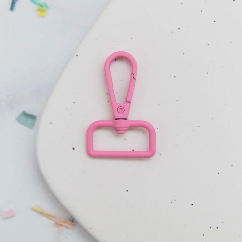 May include: A pink swivel clasp, a metal hardware component, is shown against a white background. The clasp has a rectangular base and a curved top with a spring-loaded closure. The color is a bright, solid pink.