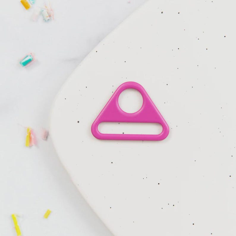May include: A bright pink, triangular-shaped metal ring with a circular hole at the top and a rectangular opening at the bottom. The ring is set against a white speckled background.