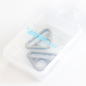 May include: Two antique bronze-colored metal triangle rings inside a clear plastic container. The rings have a circular opening on one side. The container has the text "byannie.com" printed on it in light blue.