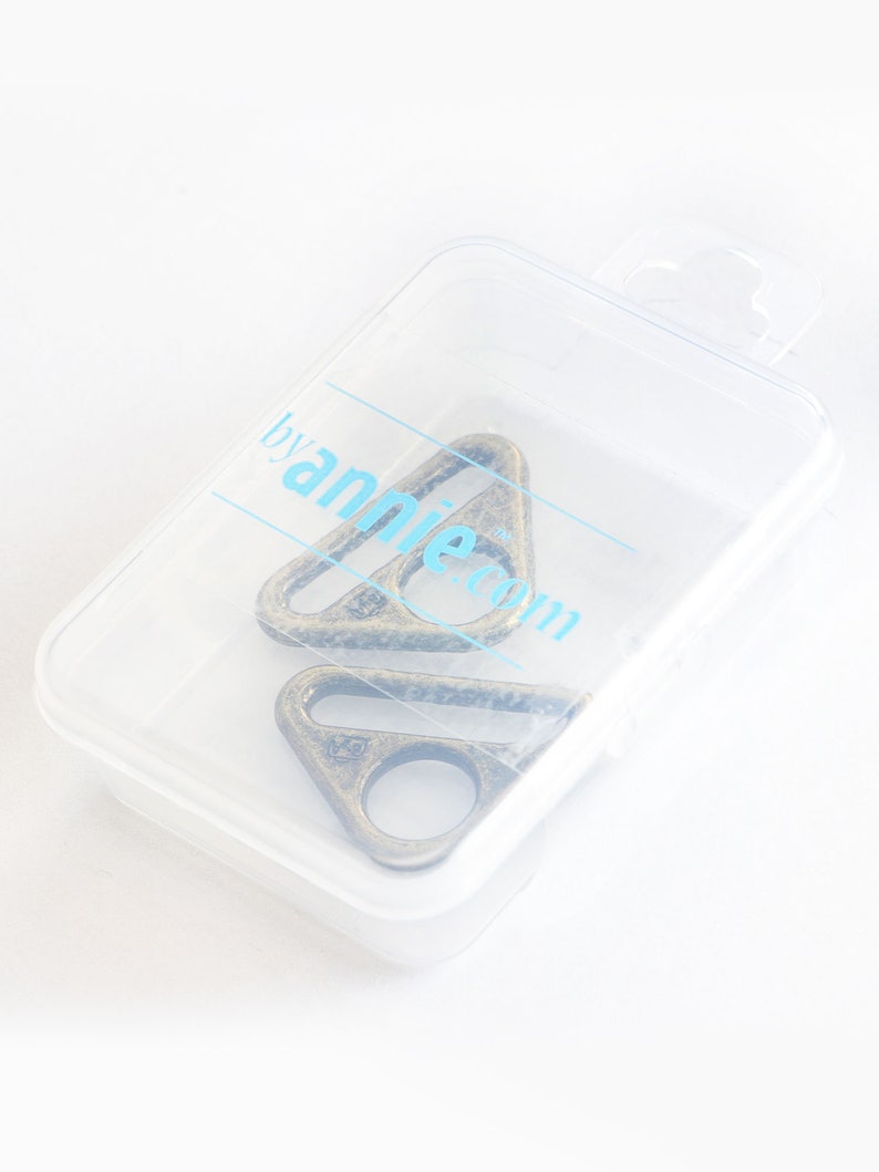 May include: Two antique bronze-colored metal triangle rings inside a clear plastic container. The rings have a circular opening on one side. The container has the text "byannie.com" printed on it in light blue.