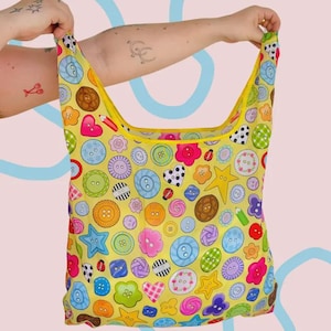 May include: A reusable shopping bag with a yellow background and a colorful button pattern. The bag has a yellow trim and handles. The buttons are various colors and shapes, including hearts, stars, and circles. The bag is held up by a person.