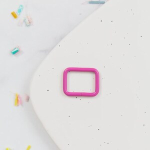 May include: A bright pink, rectangular metal ring buckle. The buckle is a vibrant magenta color and is set against a white, speckled background. The ring is a simple, square shape with rounded corners.