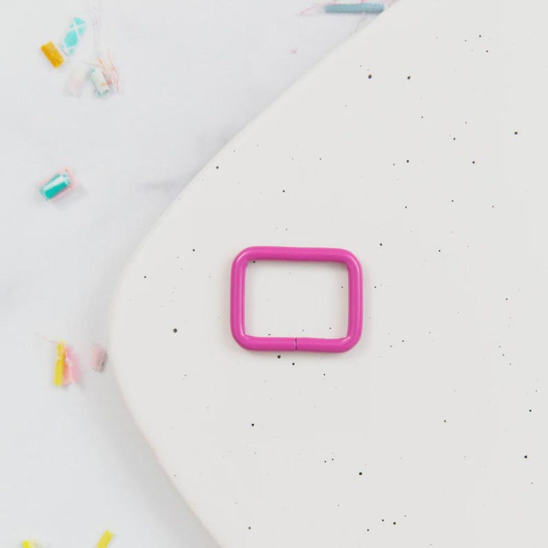 May include: A bright pink, rectangular metal ring buckle. The buckle is a vibrant magenta color and is set against a white, speckled background. The ring is a simple, square shape with rounded corners.