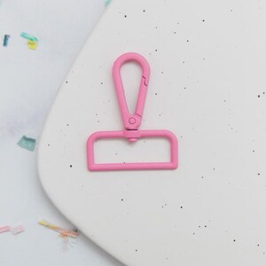 May include: A bright pink metal swivel clasp. The clasp has a rectangular base and a curved top with a spring-loaded closure. The clasp is designed to attach to a strap or other item. The background is a white surface with small black speckles.