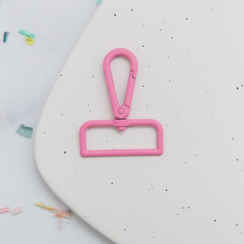 May include: A bright pink metal swivel clasp. The clasp has a rectangular base and a curved top with a spring-loaded closure. The clasp is designed to attach to a strap or other item. The background is a white surface with small black speckles.